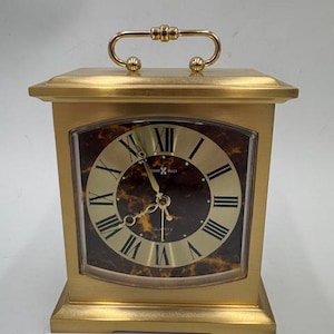 May include: A gold-colored quartz clock with a rectangular shape and a handle on top. The clock face features Roman numerals and gold hands against a brown marbled background. The clock is labeled "Howard Miller".