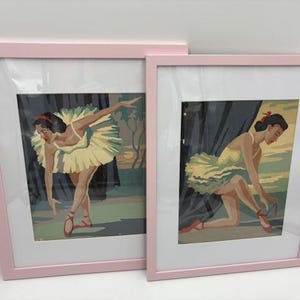 May include: Two framed paint-by-number pictures of ballerinas in pink frames. The ballerina in the first picture is posed with one arm extended. The second picture shows a ballerina kneeling and tying her shoe. The ballerinas are wearing yellow tutus.