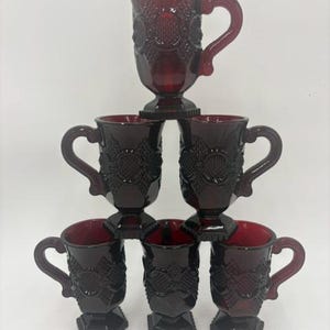 May include: A set of six ruby red glass mugs arranged in a pyramid stack. Each mug features a textured, pressed glass pattern with a circular motif, a sturdy handle, and a pedestal base. The deep red glass is translucent and uniform in color.