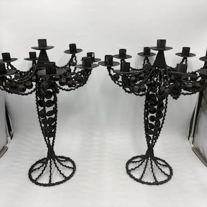 May include: Pair of black candelabras with multiple candle holders. Each candelabra has a central stem with decorative beadwork and a circular base. The candle holders are arranged in a tiered design, creating a dramatic display.