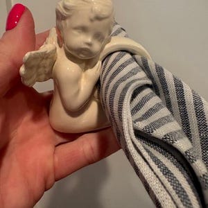 May include: A cream-colored cherub napkin ring holding a blue and white striped napkin. The cherub has detailed wings and is holding the napkin in its arms. The napkin ring is made of ceramic.