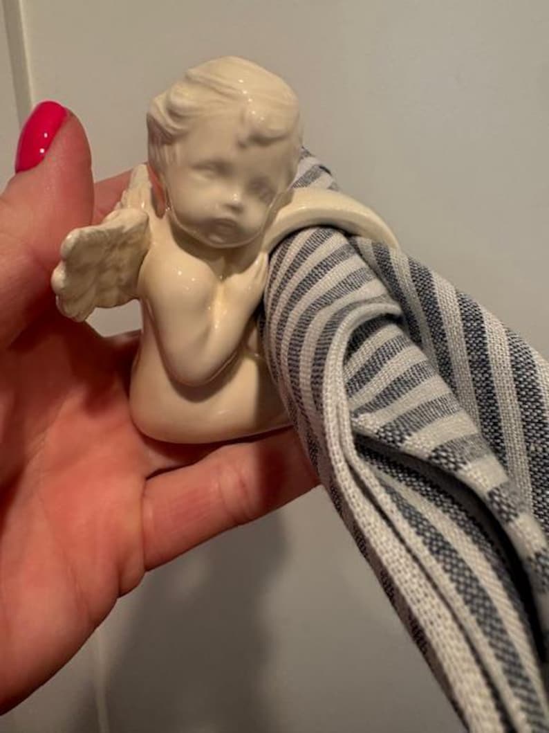 May include: A cream-colored cherub napkin ring holding a blue and white striped napkin. The cherub has detailed wings and is holding the napkin in its arms. The napkin ring is made of ceramic.