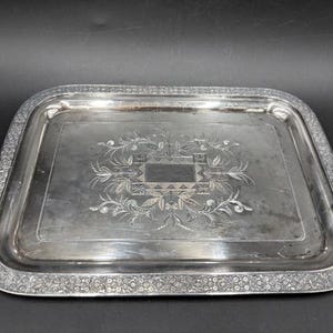 May include: A rectangular silver serving tray with a floral and geometric engraved design in the center. The tray has a detailed floral border. The tray is likely made of silver-plated metal.