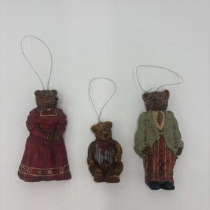 May include: Three hanging ornaments shaped like teddy bears. One bear wears a long red dress with white floral details, one is a small bear with a red bow, and one wears a light green jacket with red and white striped trousers and a tie.
