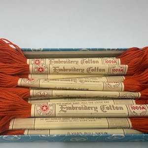 May include: A box of vintage embroidery cotton thread in a vibrant orange hue. The spools are labeled with "Embroidery Cotton" and "1009A" and are neatly arranged. The box has a blue and white patterned design.