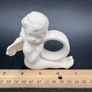May include: A cream-colored ceramic angel napkin ring. The angel is depicted in a seated position with folded wings and a ring-shaped opening for a napkin. The napkin ring measures approximately 3 inches in height.