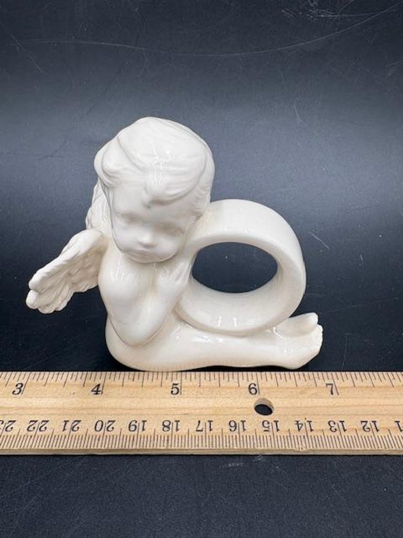 May include: A cream-colored ceramic angel napkin ring. The angel is depicted in a seated position with folded wings and a ring-shaped opening for a napkin. The napkin ring measures approximately 3 inches in height.