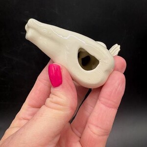 May include: A small, off-white ceramic bird whistle. The bird has a hollow body with a hole and a small wing detail. The whistle is held in a hand with pink nail polish.