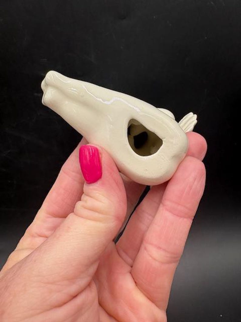 May include: A small, off-white ceramic bird whistle. The bird has a hollow body with a hole and a small wing detail. The whistle is held in a hand with pink nail polish.
