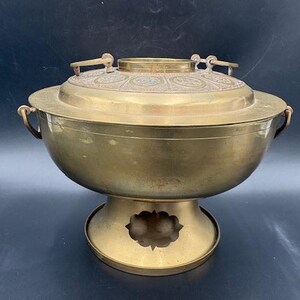 May include: A vintage brass cooking pot with a lid. The pot has a round body, a decorative lid with etched patterns, and two handles. It sits on a pedestal base with a decorative cutout. The pot has a warm, golden color.