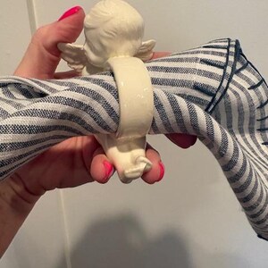 May include: A cream-colored ceramic napkin ring featuring an angel figurine. The angel has detailed wings and is holding a blue and white striped napkin. The napkin ring is a decorative item for table settings.