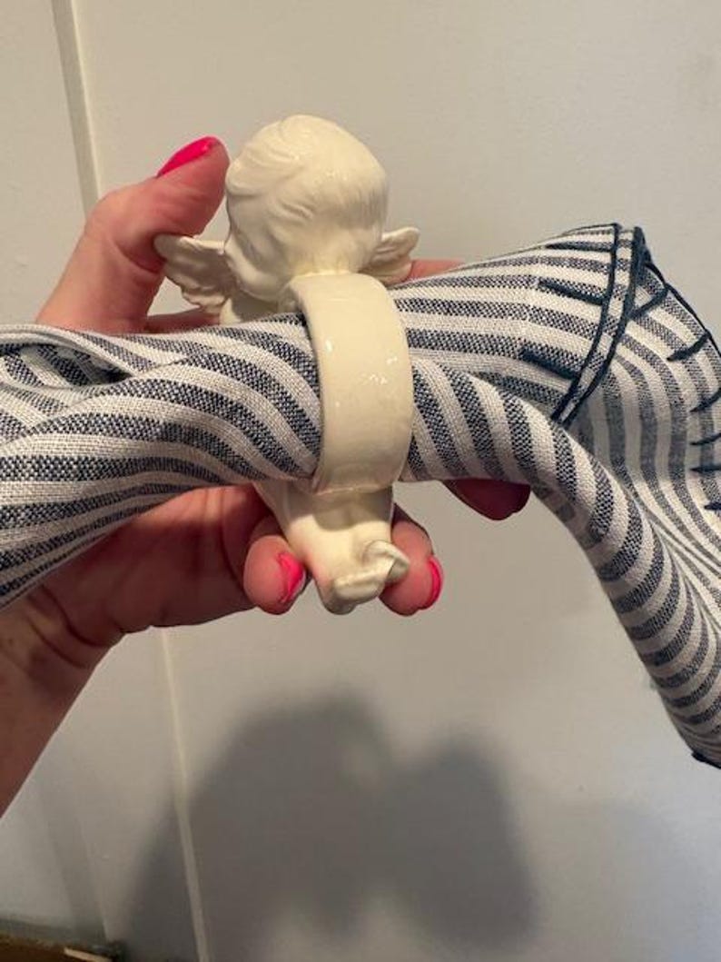 May include: A cream-colored ceramic napkin ring featuring an angel figurine. The angel has detailed wings and is holding a blue and white striped napkin. The napkin ring is a decorative item for table settings.