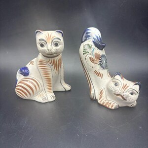 May include: Two ceramic cat figurines with a cream-colored base, featuring hand-painted brown striped patterns and blue accents. One cat sits upright, while the other is in a reclining position with its tail arched, both displaying detailed facial features.