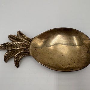 May include: A brass-colored pineapple-shaped dish. The dish has a textured surface and a detailed leaf design at the top. The bowl is oval-shaped and has a slightly worn appearance.