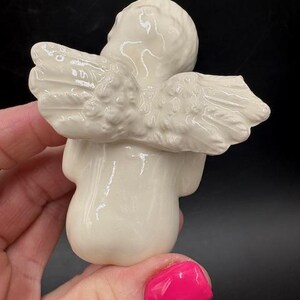 May include: A small, off-white ceramic angel figurine with detailed wings. The angel is in a seated position, with its head tilted slightly. The wings are spread out, and the figurine has a smooth, glossy finish.