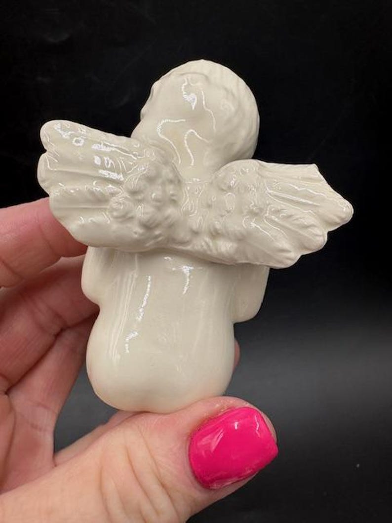 May include: A small, off-white ceramic angel figurine with detailed wings. The angel is in a seated position, with its head tilted slightly. The wings are spread out, and the figurine has a smooth, glossy finish.