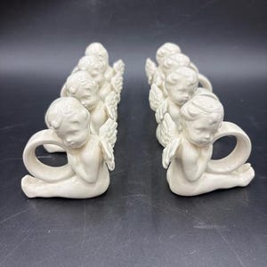 May include: Cream-colored ceramic angel napkin rings. Each angel is kneeling with folded wings and a loop for a napkin. The angels are arranged in two rows, each containing several pieces. The background is a dark, solid color.
