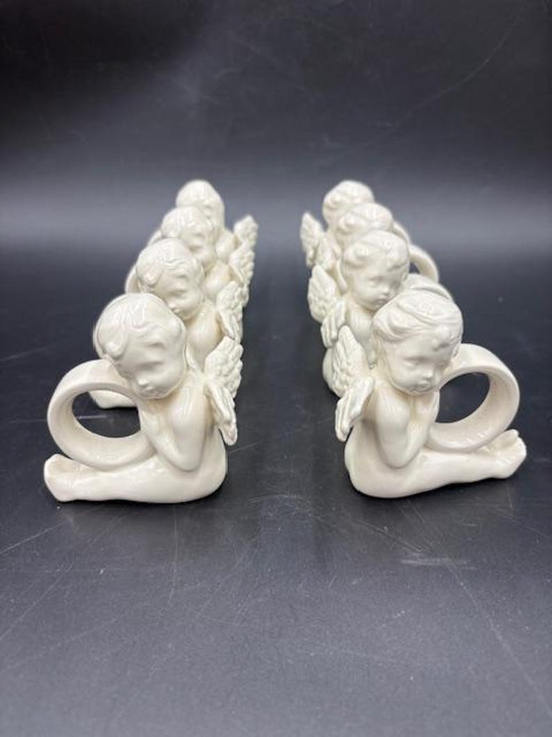 May include: Cream-colored ceramic angel napkin rings. Each angel is kneeling with folded wings and a loop for a napkin. The angels are arranged in two rows, each containing several pieces. The background is a dark, solid color.