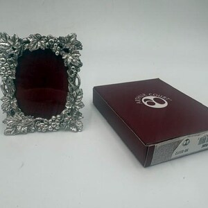 May include: A silver-toned metal photo frame with an oval opening and a deep red velvet backing, featuring a relief design of grape clusters and leaves. Beside it sits a rectangular maroon box labeled Arthur Court with a barcode.