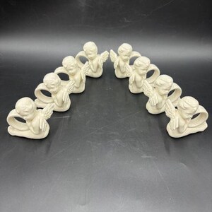 May include: A collection of nine off-white angel napkin rings. Each ring features a cherubic angel with outstretched wings and clasped hands. The angels are arranged in a semi-circle on a dark surface, creating a decorative and elegant display.