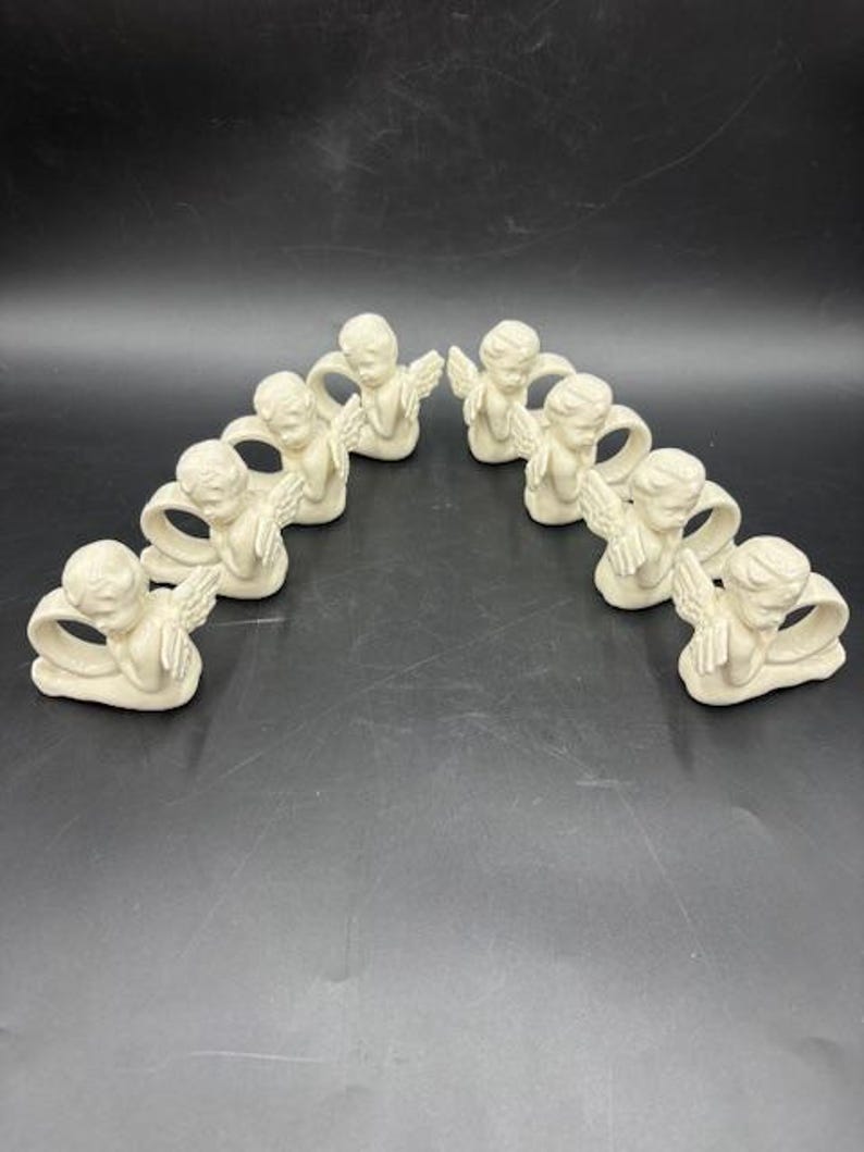 May include: A collection of nine off-white angel napkin rings. Each ring features a cherubic angel with outstretched wings and clasped hands. The angels are arranged in a semi-circle on a dark surface, creating a decorative and elegant display.