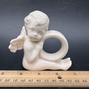 May include: A white ceramic angel napkin ring. The angel is seated with its wings spread and is holding a ring. The napkin ring is approximately 3 inches wide.