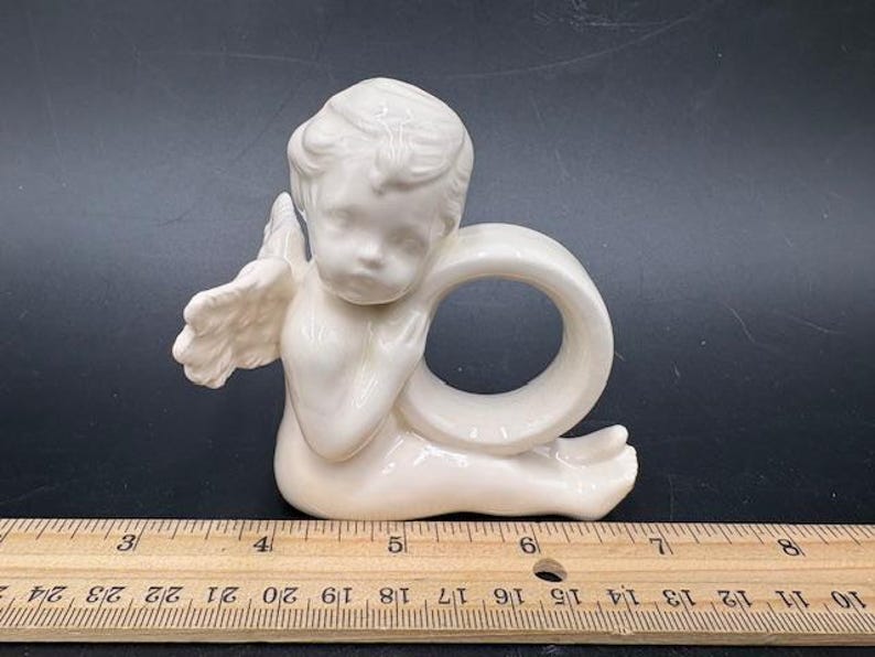 May include: A white ceramic angel napkin ring. The angel is seated with its wings spread and is holding a ring. The napkin ring is approximately 3 inches wide.