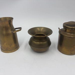 May include: Three antique brass containers. The objects include a small vase with handles, a round vase, and a milk can with a lid. The objects are arranged on a white surface.