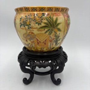 May include: A decorative ceramic bowl with an elephant motif, set on a black wooden stand. The bowl features a cream-colored background with detailed illustrations of trees, flowers, and an elephant. The stand has a carved, ornate design.