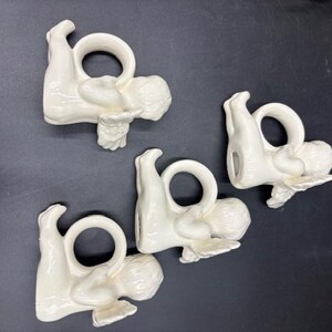 May include: Four white ceramic napkin rings, each shaped like a reclining cherub holding a ring. The cherubs are in a resting pose, with detailed wings and curly hair. The napkin rings are on a dark surface.