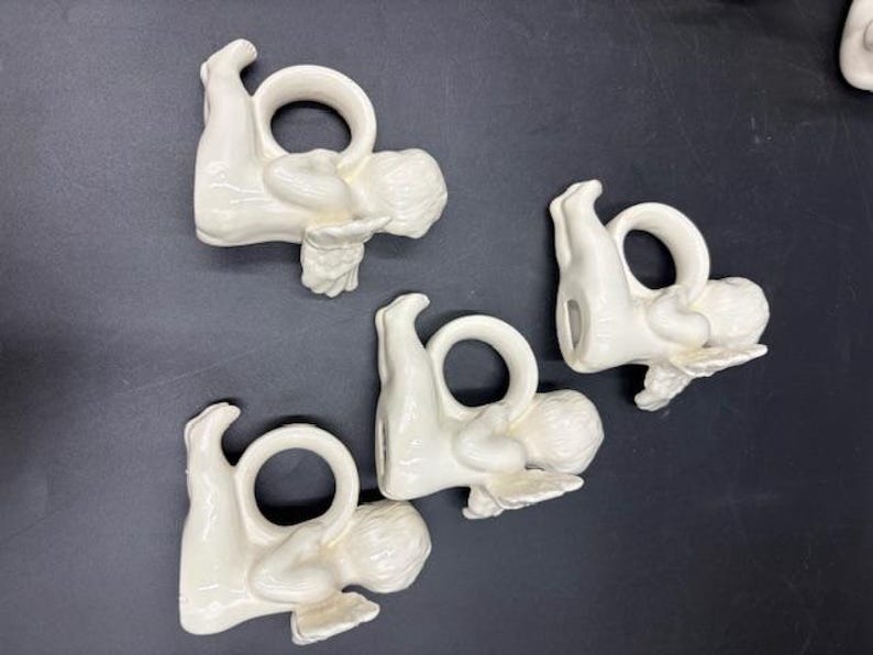 May include: Four white ceramic napkin rings, each shaped like a reclining cherub holding a ring. The cherubs are in a resting pose, with detailed wings and curly hair. The napkin rings are on a dark surface.