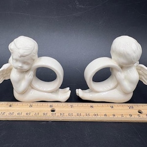 May include: Two off-white ceramic angel napkin rings. Each angel is kneeling with outstretched wings and a ring-shaped opening. The angels are positioned on a dark surface next to a wooden ruler. The ruler shows measurements in inches.