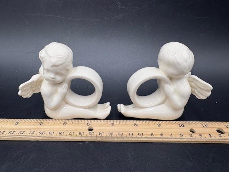 May include: Two off-white ceramic angel napkin rings. Each angel is kneeling with outstretched wings and a ring-shaped opening. The angels are positioned on a dark surface next to a wooden ruler. The ruler shows measurements in inches.