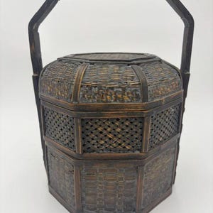 May include: A dark brown, hexagonal, multi-tiered basket with a woven bamboo texture and a tall, arched handle. The container features a lid and two stacked compartments, all constructed from dark, weathered wood-toned woven material.
