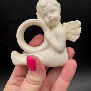 May include: Cream-colored ceramic angel napkin ring. The angel figurine has detailed wings and is holding a ring. The angel is sitting with its legs extended. The napkin ring is a decorative item.