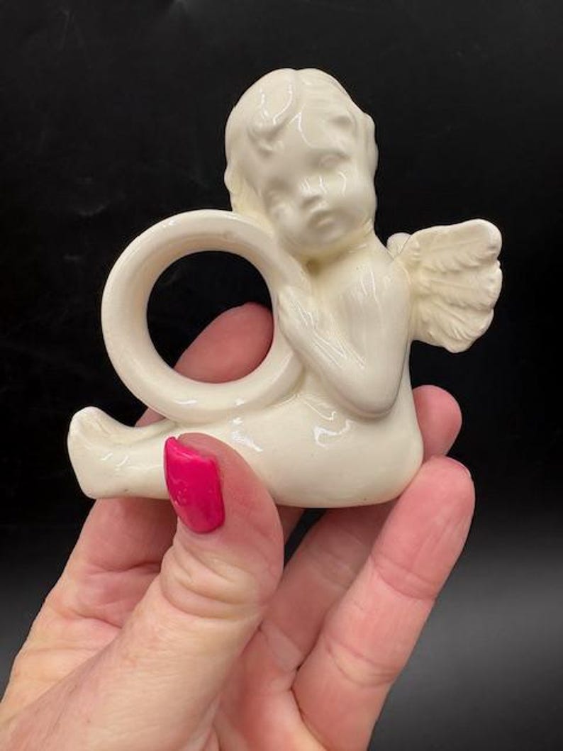 May include: Cream-colored ceramic angel napkin ring. The angel figurine has detailed wings and is holding a ring. The angel is sitting with its legs extended. The napkin ring is a decorative item.