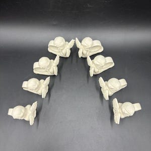 May include: A collection of eight off-white angel napkin rings arranged in an arc. Each ring features a detailed angel figure with outstretched wings. The angels are sculpted with fine details, creating a delicate and elegant appearance.