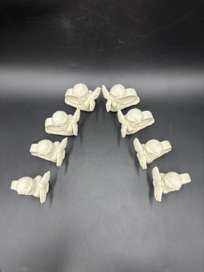 May include: A collection of eight off-white angel napkin rings arranged in an arc. Each ring features a detailed angel figure with outstretched wings. The angels are sculpted with fine details, creating a delicate and elegant appearance.