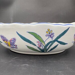 May include: White ceramic bowl with a scalloped rim and blue trim. The bowl is decorated with colorful butterflies and floral designs in shades of purple, yellow, green, and pink. A decorative piece for the home.