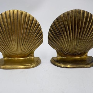 May include: Two gold-colored seashell bookends. Each bookend has a fan-shaped design with vertical lines and a flat base. The bookends are made of a metallic material and are designed to hold books upright.