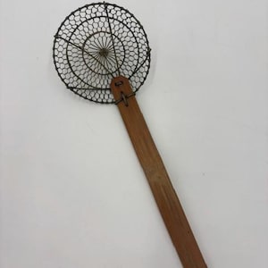 May include: A kitchen skimmer featuring a circular head made of dark metal wire mesh with a fine, intricate pattern. The head is attached to a long, flat, rectangular wooden handle by a metal fastener, creating a functional tool for straining.