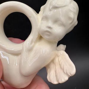 May include: Cream-colored ceramic napkin ring featuring a cherub design. The cherub has detailed facial features, wings, and is holding a small bird. The ring is smooth and has a glossy finish, suitable for decorative table settings.