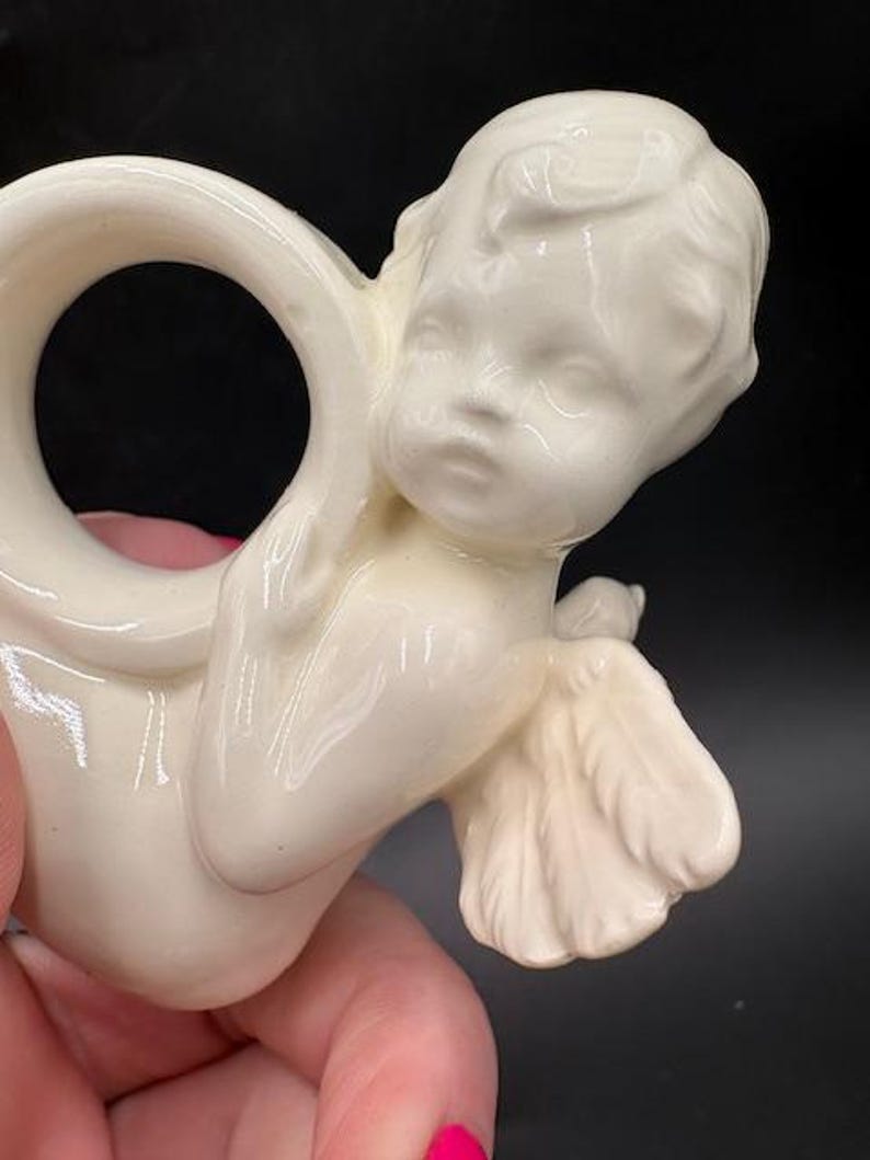 May include: Cream-colored ceramic napkin ring featuring a cherub design. The cherub has detailed facial features, wings, and is holding a small bird. The ring is smooth and has a glossy finish, suitable for decorative table settings.