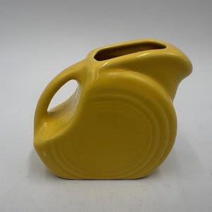 May include: A yellow ceramic pitcher with a curved handle and a spout. The pitcher has a rounded body with concentric circle detailing. The color is a bright, sunny yellow. The pitcher is likely vintage or retro.