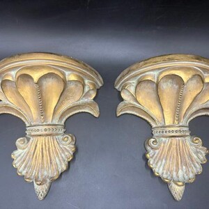 May include: A pair of wall-mounted shelves featuring a gold and weathered green finish. Each shelf has a semi-circular top, a central fluted design, and a decorative base with beaded detailing and a pointed, textured bottom element.