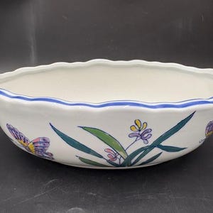 May include: White ceramic oval planter with a scalloped rim and blue trim. The planter is decorated with hand-painted floral and butterfly designs in shades of purple, yellow, and green. A decorative piece for indoor plants.
