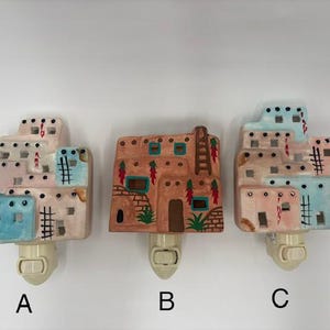 May include: Three ceramic night lights shaped like multi-level adobe buildings. Option A and C feature blue and tan tones with ladder details. Option B is tan with painted red chili peppers and green plants. Each has a white plastic plug-in base.