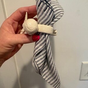 May include: A white ceramic napkin ring with a decorative design, holding a folded blue and white striped napkin. The napkin ring has a detailed, sculpted design. The napkin is folded in a decorative manner.