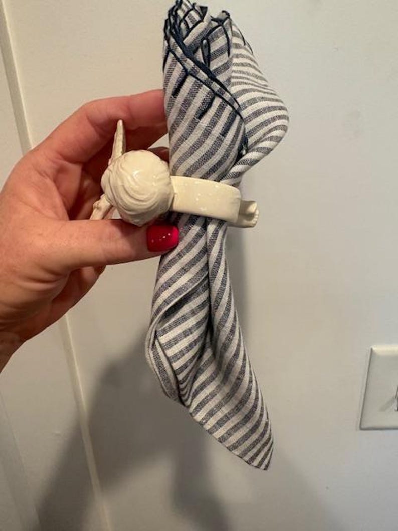 May include: A white ceramic napkin ring with a decorative design, holding a folded blue and white striped napkin. The napkin ring has a detailed, sculpted design. The napkin is folded in a decorative manner.
