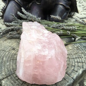 May include: A large, rough-hewn rose quartz crystal, displaying a soft pink hue. The crystal sits on a weathered, gray wooden surface, with dried lavender sprigs and a bronze statue in the background. The stone's texture is visible.
